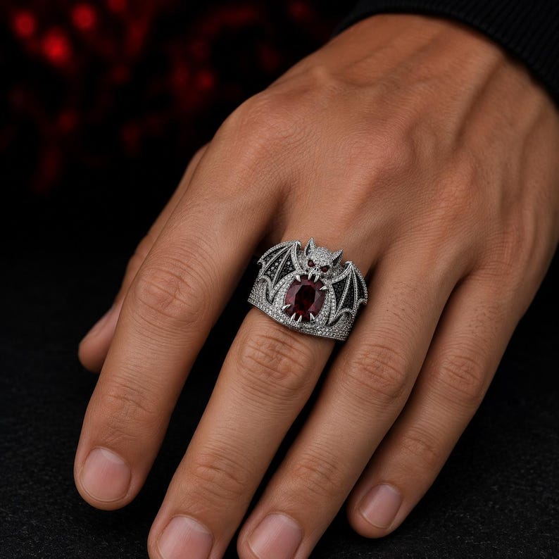 Gothic bat ring worn on finger featuring a large deep red ruby stone with black and silver vampire bat wings, fantasy gothic statement jewelry for men and women. Dark aesthetic cocktail ring with detailed pave stones, dragon inspired gothic design, Halloween jewelry, alternative fashion accessory, vampire cosplay ring, luxury gothic engagement ring, bold biker jewelry, unique handmade fantasy ring gift for gothic style lovers.
