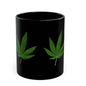 May include: Black ceramic mug with two green cannabis leaves. The leaves are detailed with visible veins and are set against a solid black background. The mug is cylindrical and appears to be empty.