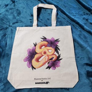 May include: Cream-colored tote bag with a colorful illustration of a snake intertwined with purple flowers. The bag has two white handles and the text "Bluestone Exotics, LLC x DAWNSNAIL" printed at the bottom.