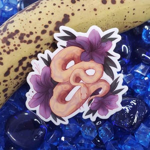 May include: A die-cut sticker featuring a coiled light orange snake surrounded by purple flowers and dark green leaves. The sticker is placed against a background of a spotted yellow banana and scattered translucent blue glass gems.