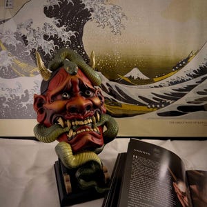 Peut inclure: Un masque Hannya rouge et marron détaillé, avec des cornes dorées et un serpent vert enroulé autour. Le masque est monté sur une base noire. En arrière-plan, une estampe de "La Grande Vague de Kanagawa."