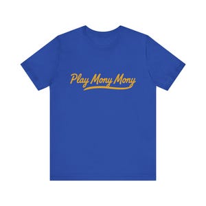 Puede incluir: Una camiseta azul real con el texto "Play Mony Mony" en amarillo, con una fuente cursiva. El texto está centrado en la parte delantera. La camiseta tiene mangas cortas y cuello redondo.