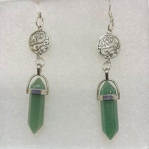 Puede incluir: Un par de pendientes colgantes con colgantes de piedras preciosas verdes. Cada pendiente tiene un disco de filigrana plateado en la parte superior, conectado a una piedra preciosa verde de forma hexagonal. Los pendientes se muestran en una caja blanca.