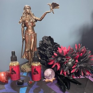 Peut inclure: Une statue en bronze d'une femme tenant un oiseau, flanquée de deux flacons à étiquettes rouges et d'une boule de cristal. Un arrangement floral noir et rouge et d'autres cristaux sont également présents. Les flacons portent le mot "MORRIGA".