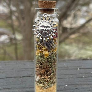 May include: A clear glass bottle filled with layers of dried herbs and flowers, topped with a cork and a silver sun charm. The bottle contains yellow, green, brown, and beige ingredients. The sun charm is detailed with a face and rays.