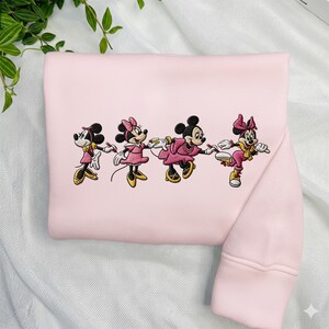 May include: A light pink sweatshirt featuring an embroidered design of four cartoon characters holding hands. The characters are dressed in pink, yellow, and black outfits. The sweatshirt has a ribbed cuff and a crew neck.
