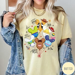 May include: A pale yellow t-shirt featuring a graphic of six character-themed balloons shaped like mouse ears. The design includes text labels for Woody, Jessie, Alien, Buzz Lightyear, Bullseye the Horse, and Bo Peep, accented with small stars and a rocket ship.
