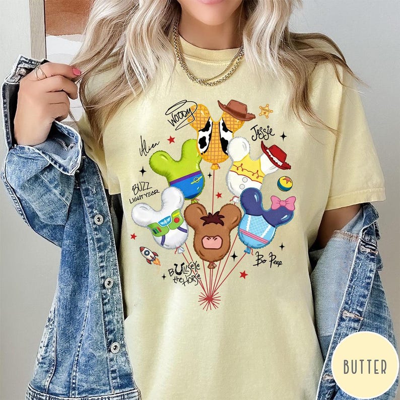 May include: A pale yellow t-shirt featuring a graphic of six character-themed balloons shaped like mouse ears. The design includes text labels for Woody, Jessie, Alien, Buzz Lightyear, Bullseye the Horse, and Bo Peep, accented with small stars and a rocket ship.