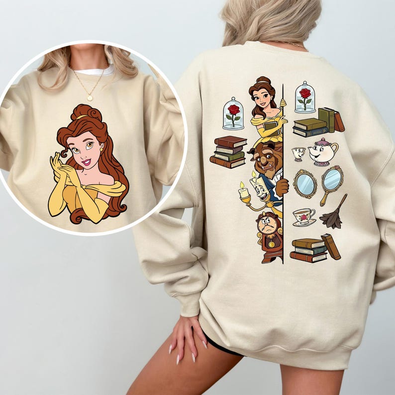 Puede incluir: Sudadera beige con un gr&aacute;fico circular de una mujer de dibujos animados con un vestido amarillo. La parte trasera de la sudadera tiene un dise&ntilde;o vertical con personajes de dibujos animados, libros y una rosa en una c&uacute;pula de cristal. El dise&ntilde;o es en tonos amarillos, marrones y azules.