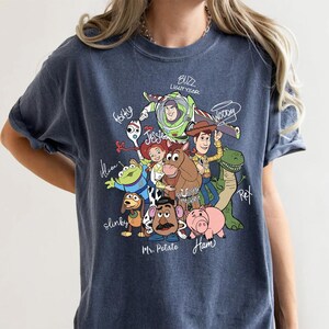 May include: A dark blue crew neck t-shirt featuring a graphic print of Toy Story characters including Buzz Lightyear, Woody, Jessie, Bullseye, Rex, Slinky, Mr. Potato Head, Ham, Forky, and an Alien, with their names written in white script.