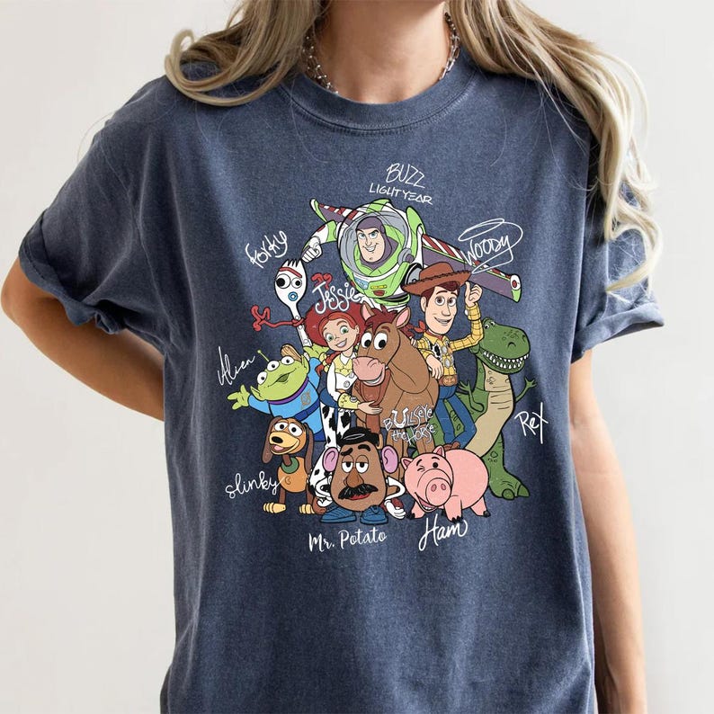 May include: A dark blue crew neck t-shirt featuring a graphic print of Toy Story characters including Buzz Lightyear, Woody, Jessie, Bullseye, Rex, Slinky, Mr. Potato Head, Ham, Forky, and an Alien, with their names written in white script.