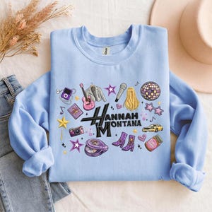 May include: Light blue sweatshirt featuring a graphic design with the text "Hannah Montana" and various illustrations. The design includes a guitar, microphone, stars, and other music-themed elements. The sweatshirt is folded on a light-colored surface.