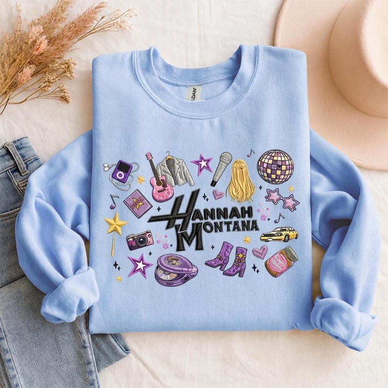 May include: Light blue sweatshirt featuring a graphic design with the text "Hannah Montana" and various illustrations. The design includes a guitar, microphone, stars, and other music-themed elements. The sweatshirt is folded on a light-colored surface.