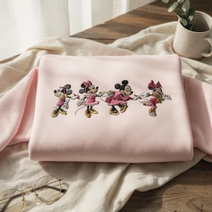 May include: A light pink sweatshirt featuring an embroidered design of four Minnie Mouse characters in various poses. The characters are dressed in pink and yellow outfits. The sweatshirt is folded on a light-colored fabric.