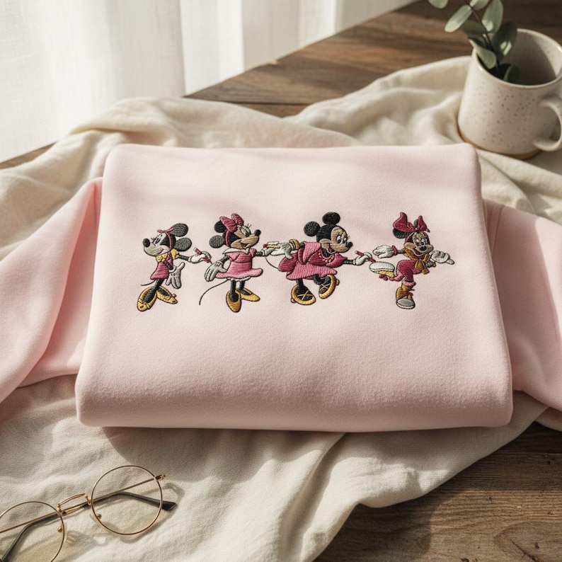 May include: A light pink sweatshirt featuring an embroidered design of four Minnie Mouse characters in various poses. The characters are dressed in pink and yellow outfits. The sweatshirt is folded on a light-colored fabric.