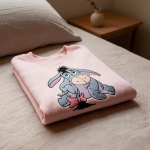 May include: A folded pale pink sweatshirt with an embroidered Eeyore design. The cartoon donkey is depicted in blue, grey, and tan, with a pink bow. The sweatshirt is neatly folded on a neutral-toned surface, likely a bed.