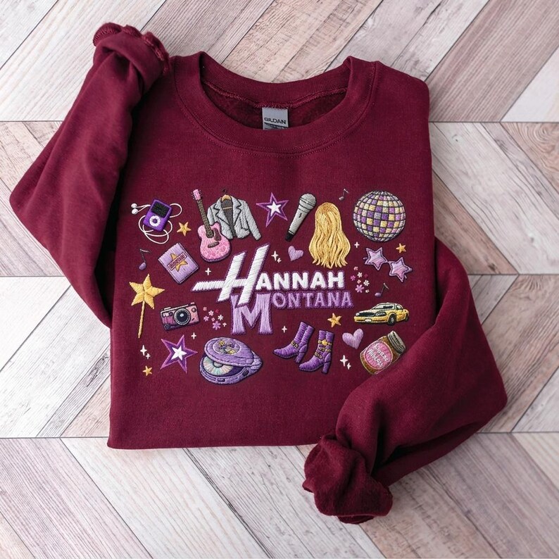 May include: Burgundy sweatshirt with embroidered Hannah Montana design. Features a guitar, microphone, disco ball, and other pop culture icons. The design includes the name "Hannah Montana" in a stylized font.