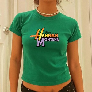 Puede incluir: Camiseta verde de manga corta con el logo de "Hannah Montana" en amarillo, naranja, morado y blanco. El logo de Disney est&aacute; encima del texto. La camiseta es de color verde liso.