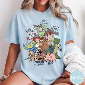 May include: A light blue t-shirt featuring a colorful graphic of Toy Story characters including Buzz Lightyear, Woody, Jessie, Bullseye, Rex, Slinky Dog, Mr. Potato Head, Ham, Forky, and an Alien, with their names written in black script around them.