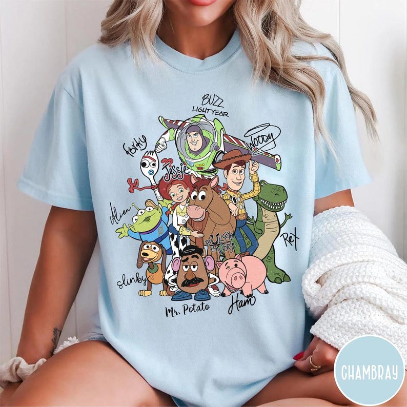 May include: A light blue t-shirt featuring a colorful graphic of Toy Story characters including Buzz Lightyear, Woody, Jessie, Bullseye, Rex, Slinky Dog, Mr. Potato Head, Ham, Forky, and an Alien, with their names written in black script around them.