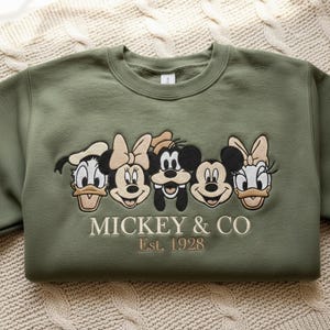 Puede incluir: Sudadera verde oliva con bordados de personajes de Disney: Pato Donald, Minnie Mouse, Mickey Mouse, Goofy y Daisy. El texto "MICKEY & CO Est. 1928" est&aacute; bordado debajo. La sudadera est&aacute; doblada sobre una superficie de punto crema.