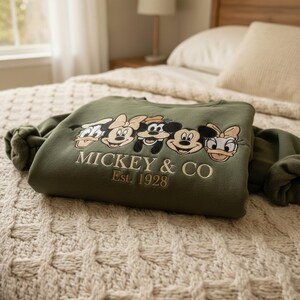 Puede incluir: Una sudadera verde oliva con im&aacute;genes bordadas de personajes de Disney: Pato Donald, Minnie Mouse, Goofy, Mickey Mouse y Daisy. El texto "MICKEY & CO Est. 1928" est&aacute; bordado debajo. La sudadera est&aacute; doblada sobre una colcha crema.