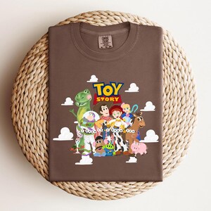 Pu&ograve; includere: Una t-shirt marrone a girocollo con una stampa grafica dei personaggi di Toy Story, tra cui Woody, Buzz Lightyear, Jessie e Rex, con il testo "TOY STORY" e "I Knew It I Knew You" circondato da illustrazioni di nuvole bianche.