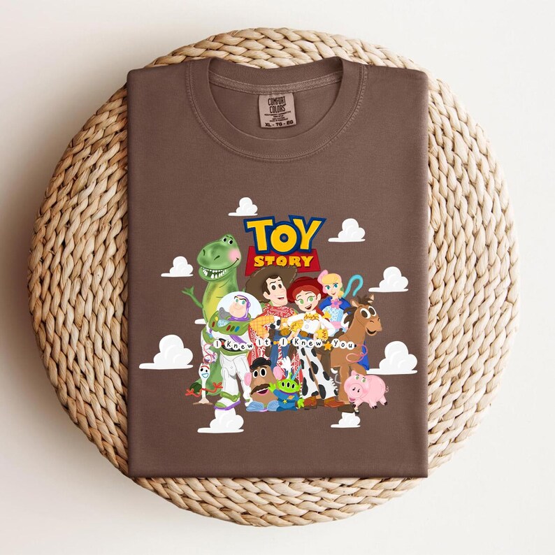 Pu&ograve; includere: Una t-shirt marrone a girocollo con una stampa grafica dei personaggi di Toy Story, tra cui Woody, Buzz Lightyear, Jessie e Rex, con il testo "TOY STORY" e "I Knew It I Knew You" circondato da illustrazioni di nuvole bianche.