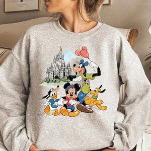 May include: A light gray sweatshirt featuring a colorful graphic of Disney characters Mickey Mouse, Donald Duck, Goofy, and Pluto. The graphic also includes a castle and red balloons. The sweatshirt is a crew neck style.