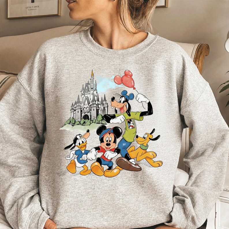 May include: A light gray sweatshirt featuring a colorful graphic of Disney characters Mickey Mouse, Donald Duck, Goofy, and Pluto. The graphic also includes a castle and red balloons. The sweatshirt is a crew neck style.