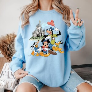 May include: Light blue sweatshirt featuring a graphic of Mickey Mouse, Donald Duck, Goofy, and Pluto in front of a castle. Goofy holds a red balloon. The sweatshirt has long sleeves and a crew neck.