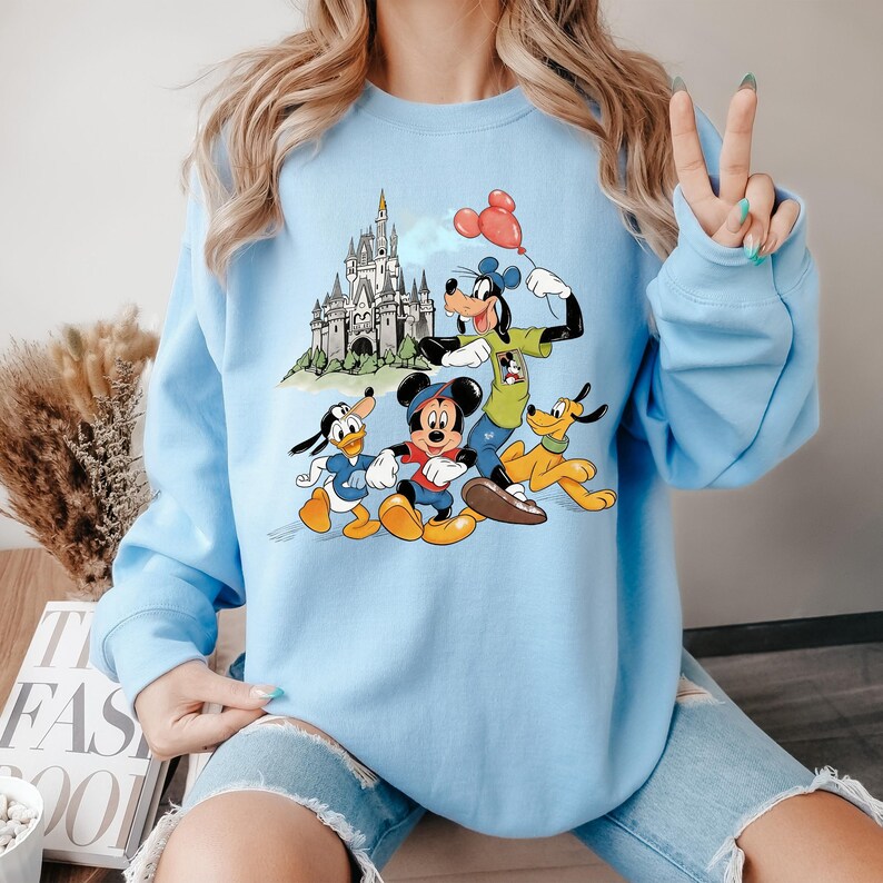 May include: Light blue sweatshirt featuring a graphic of Mickey Mouse, Donald Duck, Goofy, and Pluto in front of a castle. Goofy holds a red balloon. The sweatshirt has long sleeves and a crew neck.