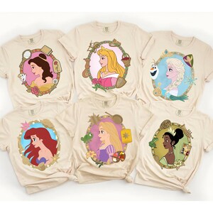 May include: Six beige t-shirts featuring colorful portraits of Disney princesses. Each shirt showcases a different princess within a decorative oval frame. The princesses include Belle, Aurora, Elsa, Ariel, Rapunzel, and Tiana.