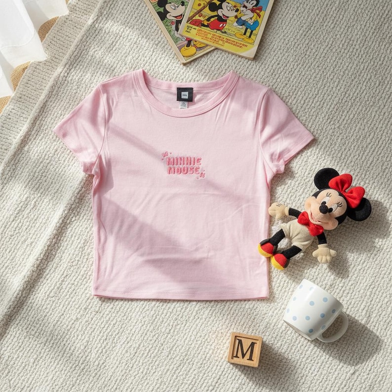 May include: A light pink t-shirt with the text "Minnie Mouse" in a cursive font. A Minnie Mouse plush toy, a polka-dot mug, and a wooden block with the letter "M" are also in the image.