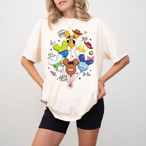May include: An off-white short-sleeve t-shirt featuring a graphic print of multiple Mickey-shaped balloons inspired by Toy Story characters. The design includes names like Woody, Jessie, Buzz Lightyear, and Bo Peep, along with small stars and a rocket.
