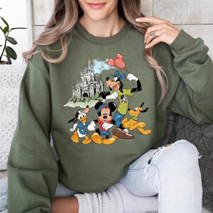 May include: Olive green sweatshirt featuring a colorful graphic of Disney characters Mickey Mouse, Donald Duck, Goofy, and Pluto in front of a castle. The characters are in their classic cartoon style, with vibrant colors and detailed designs. The sweatshirt is a comfortable, casual style.