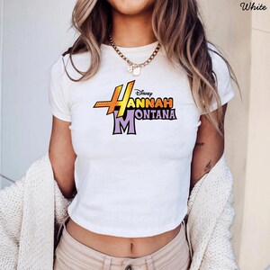 Puede incluir: Top corto blanco con el logo de Hannah Montana en naranja, amarillo y morado. El logo de Disney est&aacute; encima del nombre. La camiseta tiene mangas cortas y cuello redondo.