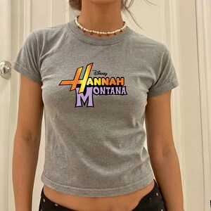 Puede incluir: Una camiseta gris de manga corta con el texto "Hannah Montana" en un dise&ntilde;o gr&aacute;fico colorido. La camiseta tiene cuello redondo y est&aacute; hecha de un material suave. Es un top corto.