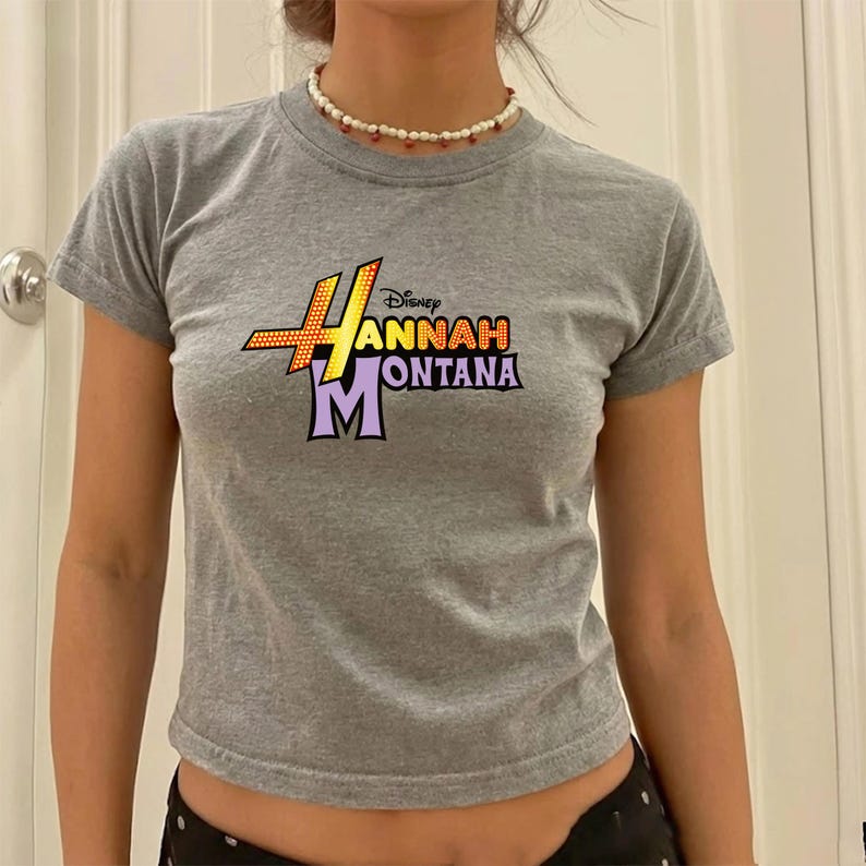 Puede incluir: Una camiseta gris de manga corta con el texto "Hannah Montana" en un dise&ntilde;o gr&aacute;fico colorido. La camiseta tiene cuello redondo y est&aacute; hecha de un material suave. Es un top corto.