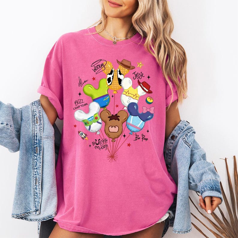 May include: A bright pink t-shirt featuring a graphic of six balloons shaped like mouse ears, each themed after a different character. The balloons include designs inspired by Woody, Jessie, Buzz Lightyear, Bo Peep, Bullseye, and an Alien, with names written in script.