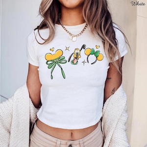 May include: White crop top with cartoon graphics of a yellow bow, Pluto, and Mickey Mouse ears. The shirt has a crew neck and short sleeves. The word "White" is printed in the upper right corner.