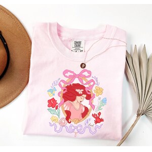 May include: A pale pink t-shirt with a framed illustration of a red-haired character, surrounded by flowers, a crab, and fish. The shirt has a crew neck and short sleeves. A gold necklace with a pendant is draped across the shirt.