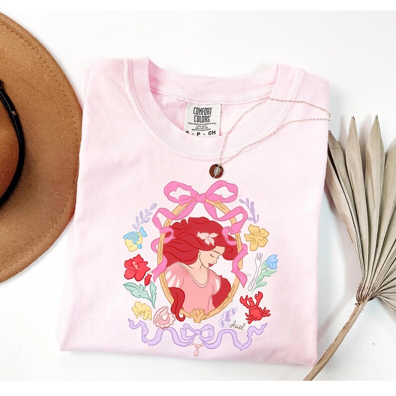 May include: A pale pink t-shirt with a framed illustration of a red-haired character, surrounded by flowers, a crab, and fish. The shirt has a crew neck and short sleeves. A gold necklace with a pendant is draped across the shirt.