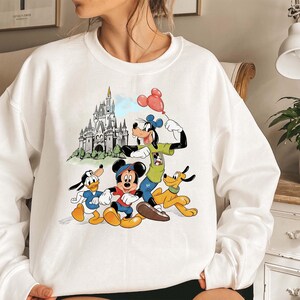 May include: White sweatshirt featuring a colorful graphic of Disney characters Mickey Mouse, Donald Duck, Goofy, and Pluto. The design includes a castle and a red balloon. The sweatshirt is a crew neck style.