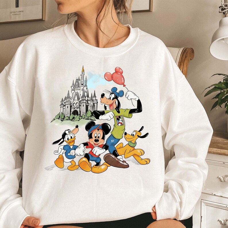 May include: White sweatshirt featuring a colorful graphic of Disney characters Mickey Mouse, Donald Duck, Goofy, and Pluto. The design includes a castle and a red balloon. The sweatshirt is a crew neck style.