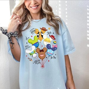 May include: A light blue t-shirt featuring a graphic print of multiple balloons shaped like mouse ears, each decorated to resemble characters including Woody, Jessie, Buzz Lightyear, and Bo Peep, with their names written in script text around the balloons.