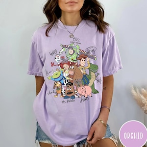 May include: A light purple short-sleeve t-shirt featuring a graphic print of Toy Story characters including Buzz Lightyear, Woody, Jessie, Forky, Rex, Slinky, Mr. Potato Head, Ham, and an Alien, with their names written in black cursive script.