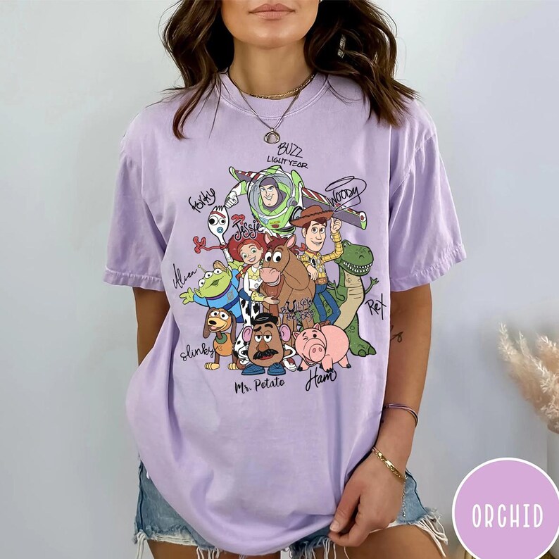 May include: A light purple short-sleeve t-shirt featuring a graphic print of Toy Story characters including Buzz Lightyear, Woody, Jessie, Forky, Rex, Slinky, Mr. Potato Head, Ham, and an Alien, with their names written in black cursive script.