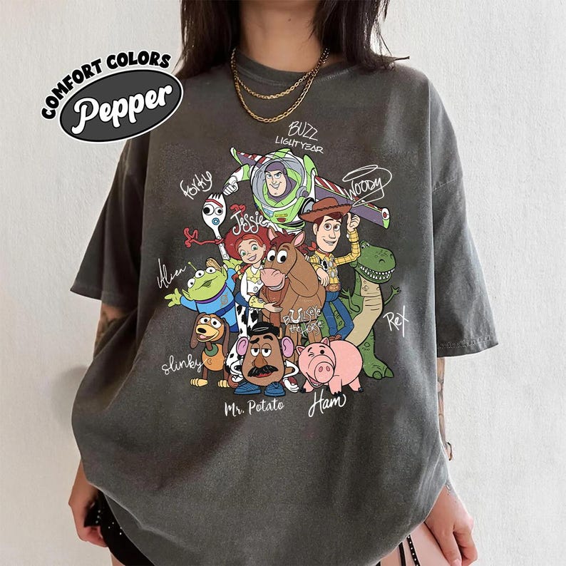 May include: A charcoal gray t-shirt featuring a graphic print of Toy Story characters including Buzz Lightyear, Woody, Jessie, Bullseye, Rex, Slinky Dog, Mr. Potato Head, Hamm, Forky, and an Alien, with character names written in white script text.