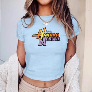 Puede incluir: Top corto azul claro con el texto "Hannah Montana" en un dise&ntilde;o colorido y estilizado. El logotipo de Disney est&aacute; encima del texto. La camiseta tiene mangas cortas y cuello redondo.